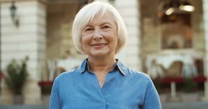 Close up of Caucasian joyful pretty gray-haired old woman in blue jeans shirt smiling outdoors. Happy good looking grandmother with gray hair laughing. Portrait of senior beautiful lady smile.