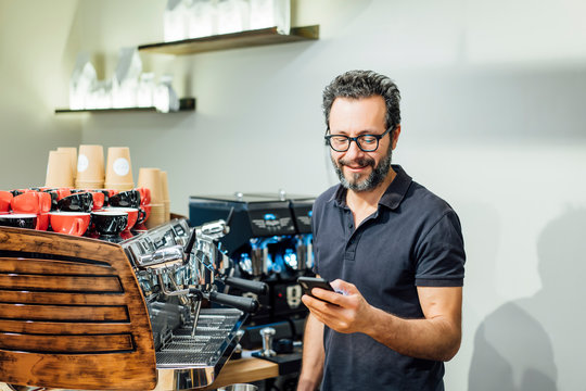 Male Barista Working In The Cafeteria Using Mobile Phone.