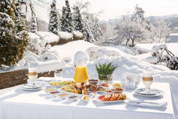 Fototapeta premium Winter Breakfast pancakes on the terrace outside the restaurant on the background of snow. Pancakes, fruit, juice, and coffee