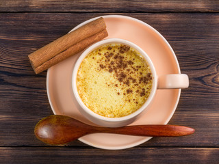 Top view of a cup of golden milk with wooden spoon and cinnamon on a wooden background