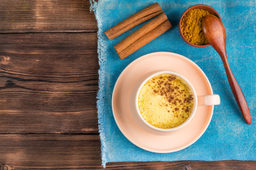 Top view of a cup of golden milk with turmeric and cinnamon and wooden spoon on a blue background. With copy space.
