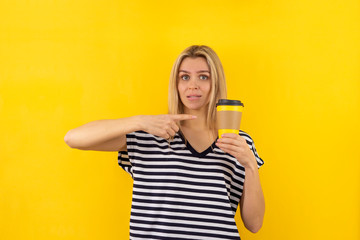 A young woman in striped t-shirt holds and pointing with finger on a disposable yellow coffee cup on a yellow background. Copy space on a cup