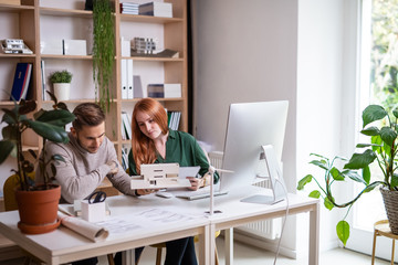 Architects with model of house sitting at the desk indoors in office, working.