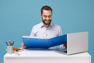 Smiling young bearded man in light shirt work at white desk with pc laptop isolated on pastel blue background. Achievement business career concept. Mock up copy space. Hold folder for papers document.