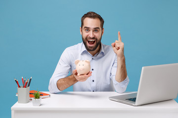 Excited young man in light shirt work at desk with pc laptop isolated on pastel blue background. Achievement business career concept. Hold piggy money bank, point index finger up with great new idea.