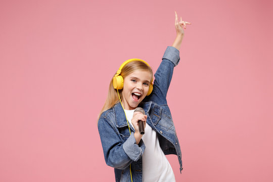 Cute little kid girl 12-13 years old isolated on pastel pink background. Childhood lifestyle concept. Mock up copy space. Listen music with headphones sing song in microphone showing horns up gesture.