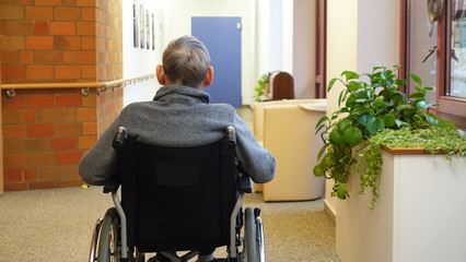 An elderly man, senior, is sitting in a wheelchair in the corridor near the window with his back to...
