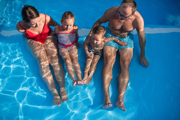 Young family with two small children sitting in swimming pool outdoors.