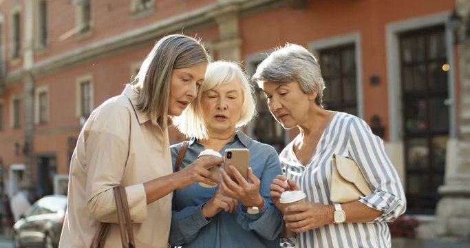 Three beautiful and cheerful Caucasian women with coffee to-go in hands laughing while scrolling photos on smartphone outdoor. Happy joyful grandmothers smiling while watching pictures on smartphone.