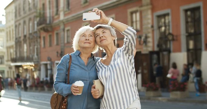 Two Caucasian beautiful old women smiling while taking selfie photo with smartphone camera on street in city center. Senior happy ladies posing to phone while taking selfies picture.
