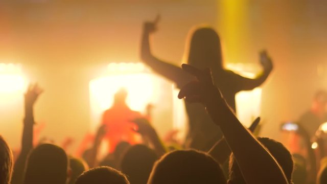 Super slow motion: people crowd silhouettes partying, cheering and raising hands up at rock concert in front of stage of nightclub. Bright colorful stage lighting. Nightlife and entertainment concept