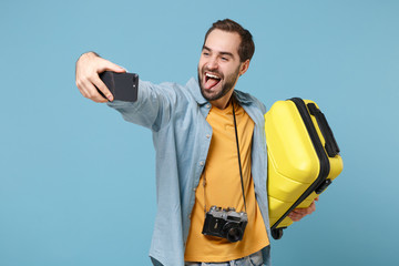 Crazy traveler tourist man in yellow clothes with photo camera isolated on blue background. Passenger traveling abroad on weekends. Air flight journey. Hold suitcase doing selfie shot on mobile phone.