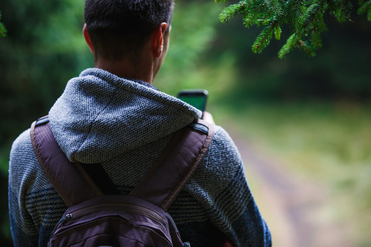 A Young Man With A Backpack In The Woods Looking At The Phone-a View From The Back. Orientation On A Walk Along A Forest Path Using The Navigator In Your Phone