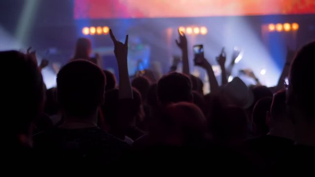 Nightlife and entertainment concept. People crowd partying, raising hands up, showing sign - devil's horns gesture at rock concert in front of stage of nightclub with flashing light: super slow motion