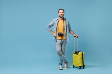 Smiling traveler tourist man in yellow summer casual clothes with photo camera isolated on blue background. Male passenger traveling abroad on weekends. Air flight journey concept. Holding suitcase.