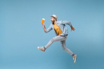 Side view of traveler tourist man in summer clothes isolated on blue background. Male passenger traveling abroad on weekend. Air flight journey concept. Jumping like running, holding passport ticket.