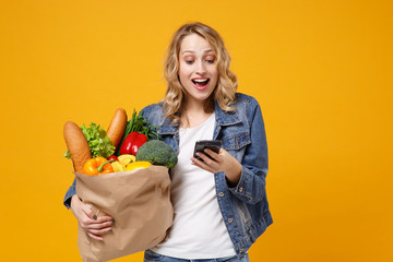 Cheerful young woman in denim clothes isolated on orange background. Delivery service from shop or restaurant concept. Hold brown craft paper bag for takeaway mock up with food product mobile phone.
