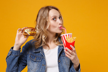 Pretty young woman in denim clothes isolated on orange background. Proper nutrition or American classic fast food concept. Mock up copy space. Hold burger french fries potatoes drink cola or soda.