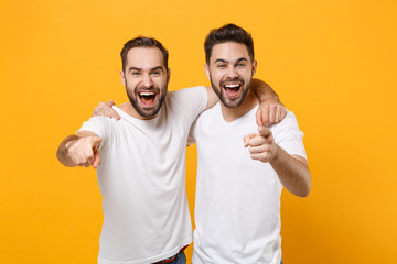 Laughing young men guys friends in white blank empty t-shirts posing isolated on yellow orange background studio portrait. People lifestyle concept. Mock up copy space. Point index fingers on camera. - obrazy, fototapety, plakaty
