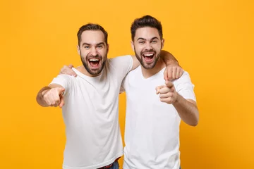 Fotobehang Laughing young men guys friends in white blank empty t-shirts posing isolated on yellow orange background studio portrait. People lifestyle concept. Mock up copy space. Point index fingers on camera. © ViDi Studio