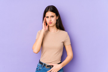 Young caucasian woman isolated on purple background blows cheeks, has tired expression. Facial expression concept.