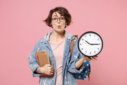 Amazed young woman student in denim clothes eyeglasses, backpack posing isolated on pastel pink background. Education in high school university college concept. Mock up copy space. Hold books clock.