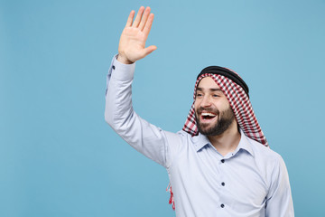 Naklejka premium Joyful bearded arabian muslim man in keffiyeh kafiya ring igal agal casual clothes isolated on pastel blue background. People religious lifestyle concept. Waving greeting with hand as notices someone.