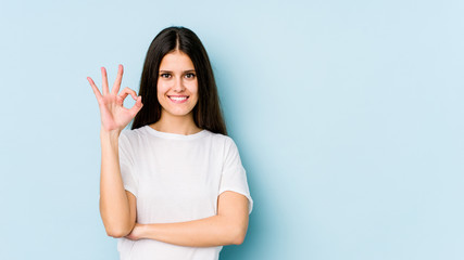 Young caucasian woman isolated on blue background winks an eye and holds an okay gesture with hand.