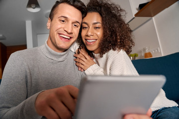Romantic Date. Young multiethnic couple at home sitting on sofa using digital tablet together laughing joyful close-up