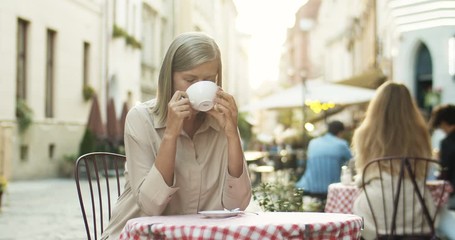 Good looking senior Caucasian smiled woman with gray hair sipping coffee while sitting at table in cafe terrace outdoors. Lady on retirement drinking tea and resting. Pensioner spending time nice. - Powered by Adobe