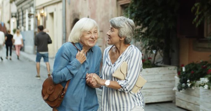 Two Caucasian pretty female pensioners with gray hait and in nice outfits walking the city street and talking joyfully. Beautiful friends women stepping outdoor and sharing secrets or gossips.