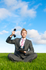 Calm businessman meditating in bright green meadow holding up the receiver of an old-fashioned...
