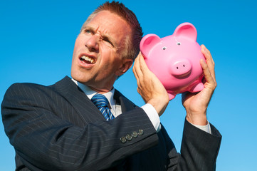 Frustrated businessman shaking a pink piggy bank listening for coins outdoors in bright blue sky