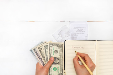 Expenses planning concept. Top view of receipts, woman holding pen and cash and writing words Expenses in a notepad on white wooden table. Image with copy space, flat lay