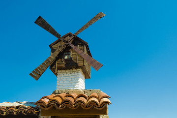 Obraz premium Beautiful wooden old small mill on a blue clear sky in a clear sunny day. Decorative traditional white mill with a brown roof on the tiled roofing of a house on an ancient street of a European city.