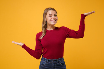 Smiling young blonde woman girl in casual clothes posing isolated on yellow orange background. People lifestyle concept. Mock up copy space. Spreading pointing hands aside, holding something in palms.