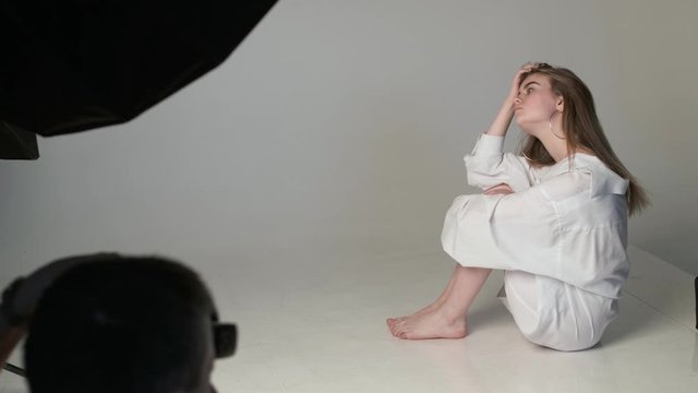 backstage of male photographer taking pictures of teen caucasian girl with long hair posing in white shirt on studio floor with bare feet. model tests of young pretty girl. photo shooting on cyclorama