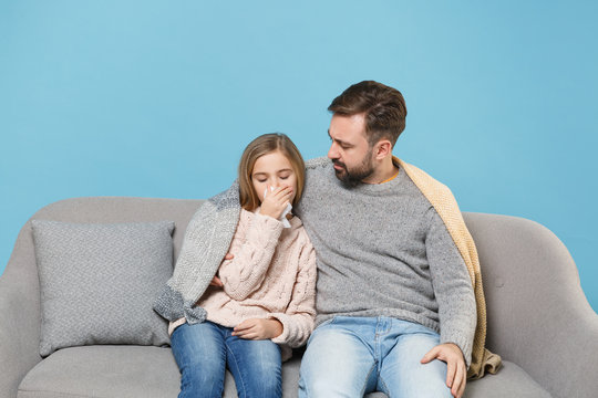 Man in knitted sweater with sick child baby girl. Father little daughter isolated on blue background. Love family parenthood childhood concept. Sit on couch, having runny nose, blowing nose to napkin.