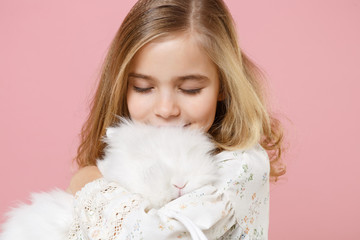 Little pretty blonde kid girl 11-12 years old in light spring dress hold fluffy white bunny rabbit isolated on pastel pink background children studio portrait. Childhood lifestyle Happy Easter concept