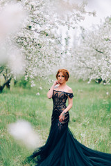 A young woman in a black dress is standing in a blooming garden. Full-length portrait.