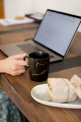 The girlÕs hand is holding on to a black mug in the japanese kintsugi style and next to it is a sandwich for a snack on a white plate. On a wooden table is a laptop computer. Hot drink tea or coffee.