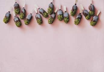 Ice cream popsicle pattern. Flat-lay of chocolate glazed ice cream pops with pistachio nuts icing over pastel pink background, top view, copy space. Summer cold sweet healthy vegan dessert