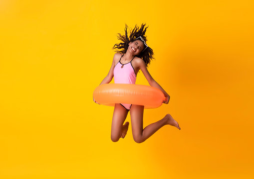 Happy young woman dressed in swimwear with rubber ring and jumping on yellow background.