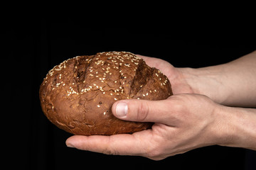 Man's hands hold tasty fresh loaf of dark bread with sesame seeds on black background