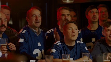 MED Model released, American football fans wearing generic blue uniform watching a game on a TV in a sport pub, rooting for their team. 4K UHD, shot on ARRI Alexa Mini with Cooke S4 lenses