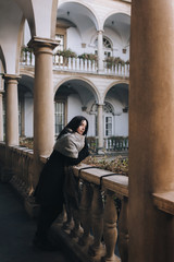 A young caucasian black-haired girl in a dark autumn coat, boots and a wide scarf-wrap stands on the balcony with a gray colonnade and looks down thoughtfully. Lviv, Italian courtyard, Ukraine.