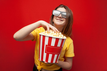 beautiful young girl in 3D glasses eating popcorn and watching a movie on a red color background