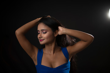 Close up fashion portrait of an young and attractive Indian Bengali brunette girl with blue western dress in front of a black studio background. Indian fashion portrait and lifestyle.