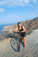 Attractive and smiling young Latina, dressed in black, riding a mountain bike on a bright and sunny summer day on a mountain and at the bottom the ocean. healthy lifestyle