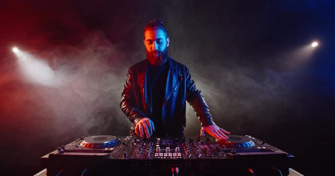 Cool bearded disc jockey working at mixer controller in a nightclub. Authentic dj performing in neon lights and smoke - nightlife concept 4k footage